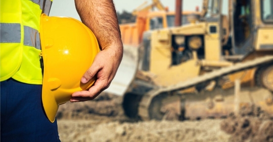 Construction worker holding a hard hat with construction equipment in the background.